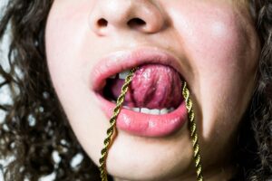 Close-up of a person's open mouth with a gold rope chain draped over their tongue.