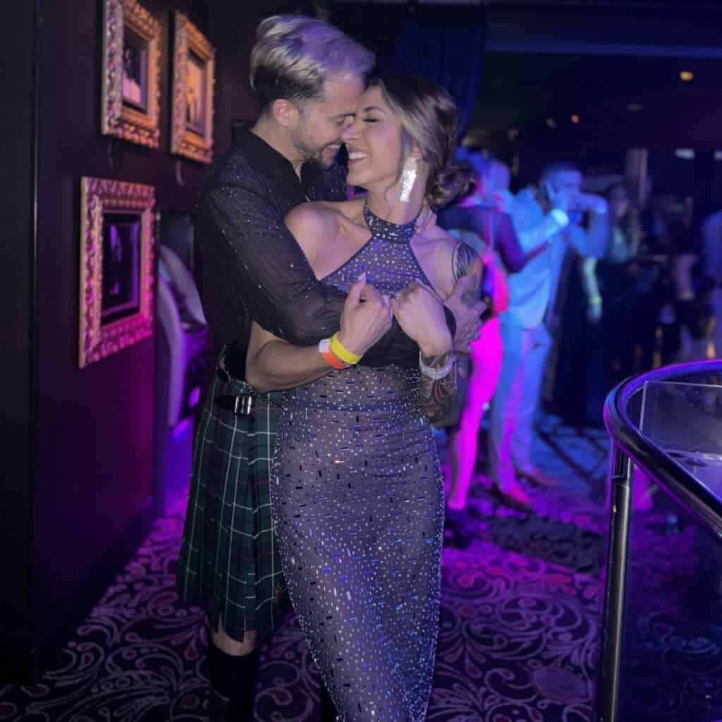 Couple dancing at a party with colorful lighting and framed artwork on the wall.